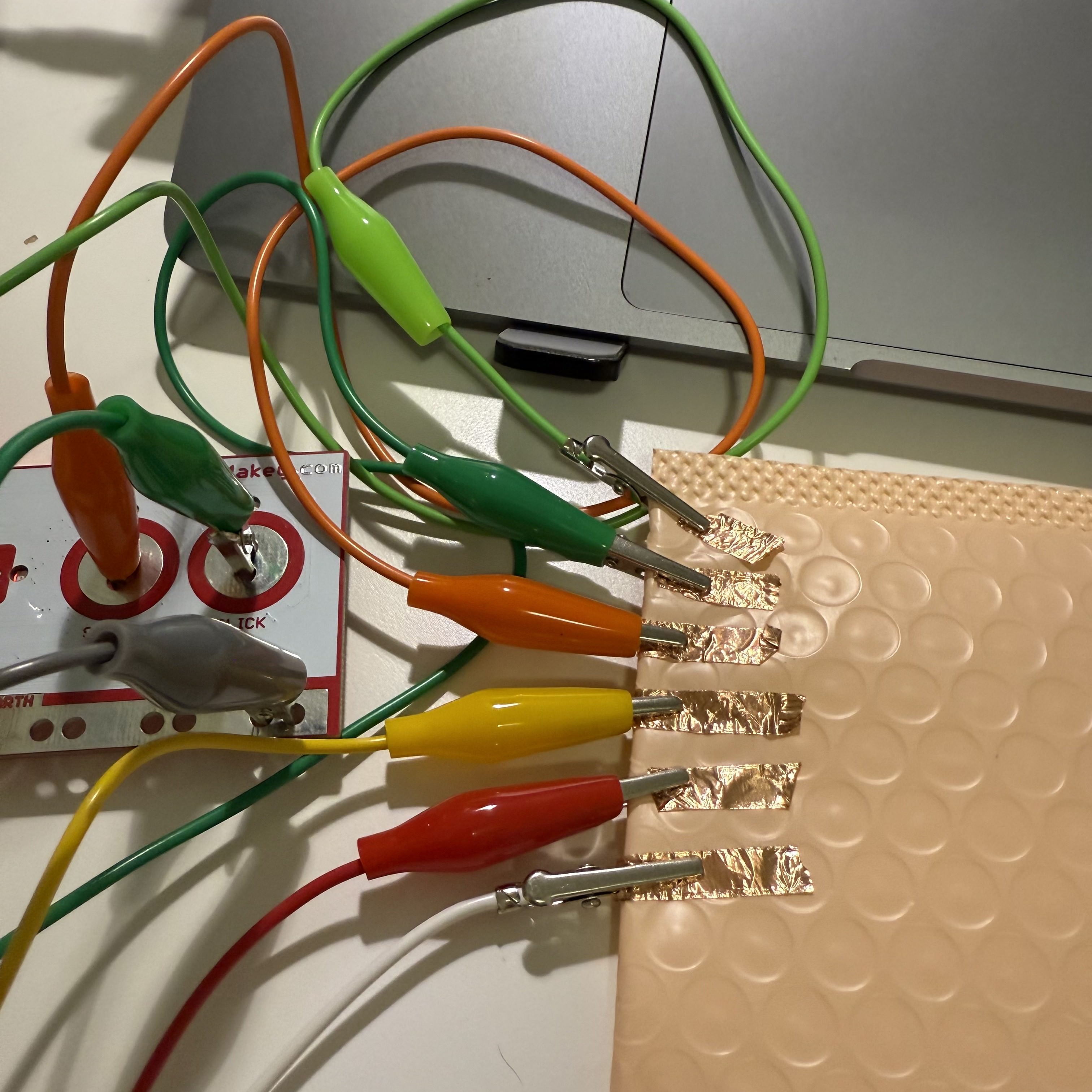 The Makey Makey board is seen with many colorful wires connected to it. Each wire is clipped to a copper strip that is attached to a bubbly textured pad. This setup allows me to interact with my computer in a unique way by touching these copper contacts.